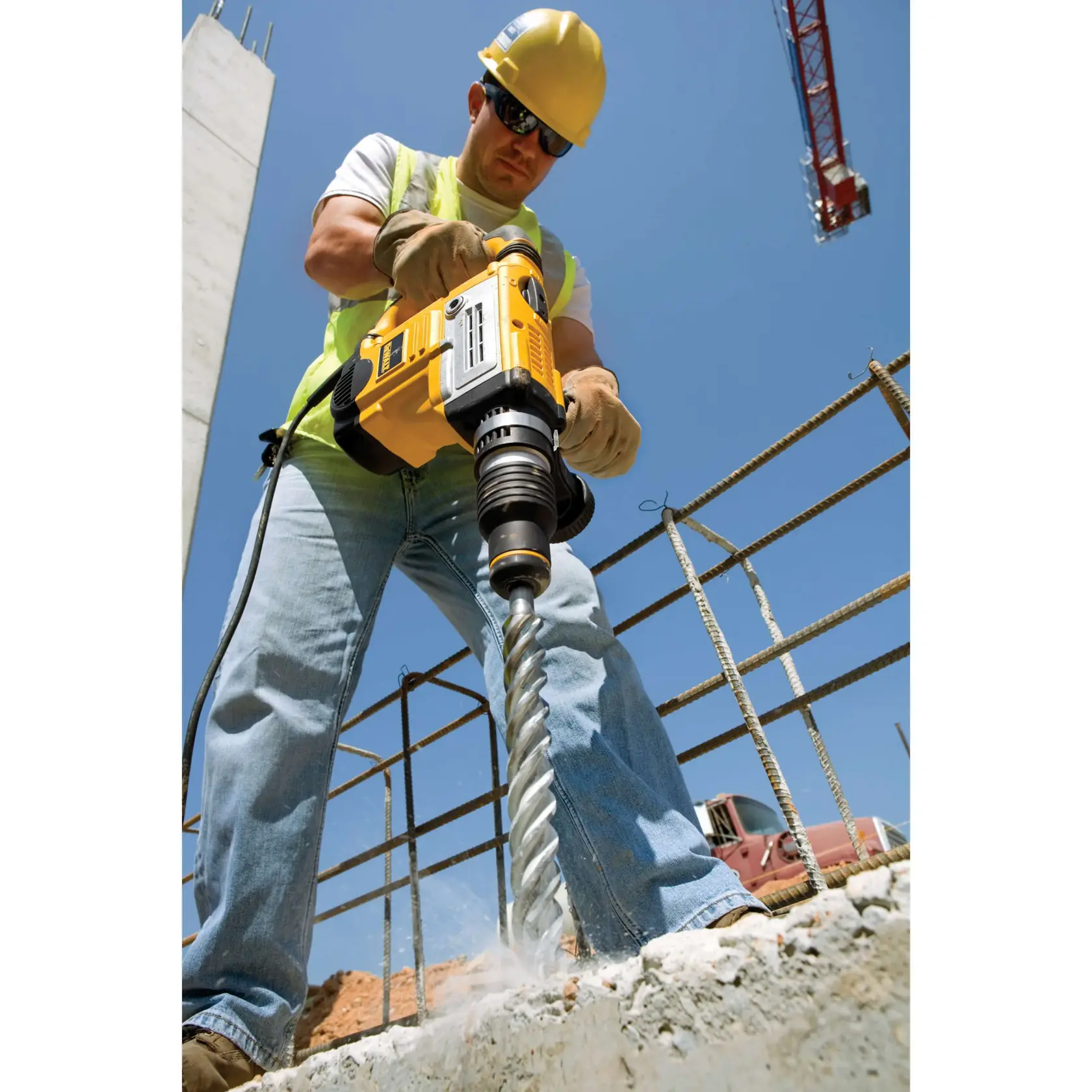 Person drilling concrete with a DEWALT rotary hammer at a construction site. Safety gear and crane in background.