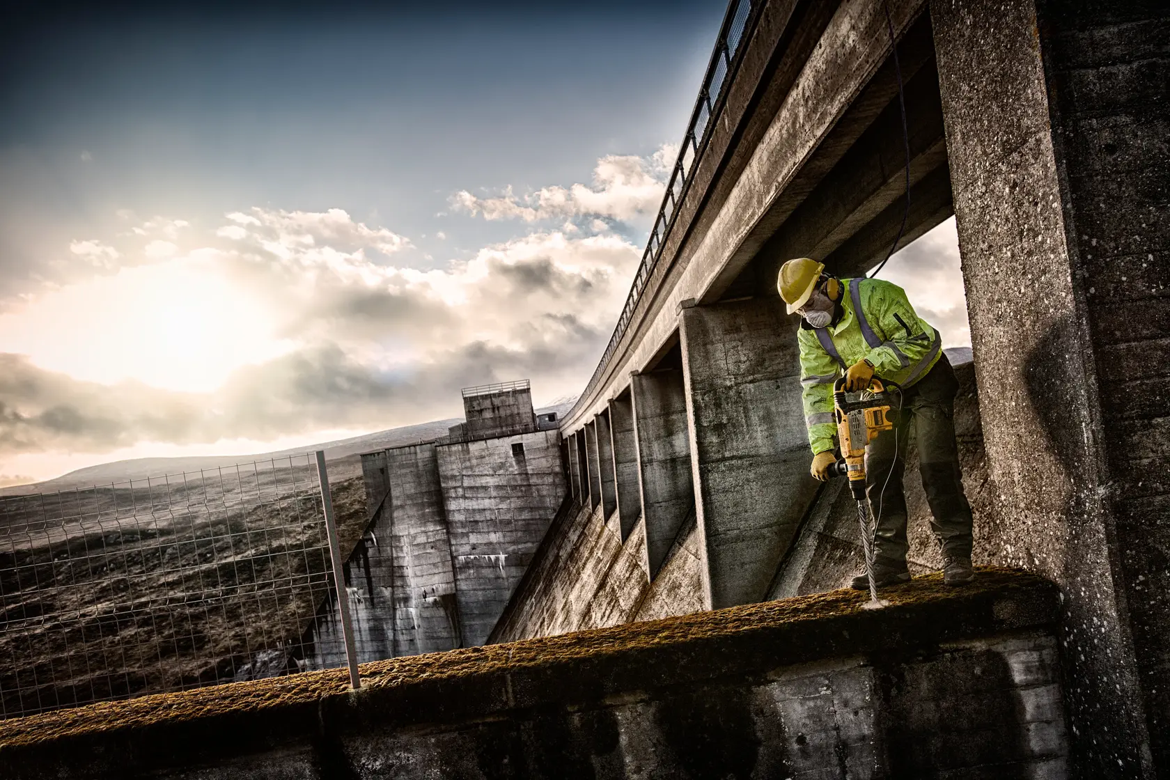 Worker with DEWALT tool drilling concrete on a dam structure outdoors at sunrise.