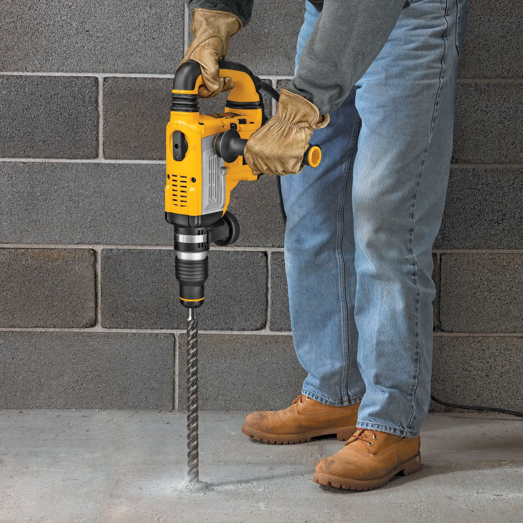 Person drills concrete floor with a yellow DEWALT rotary hammer drill, wearing gloves and boots.