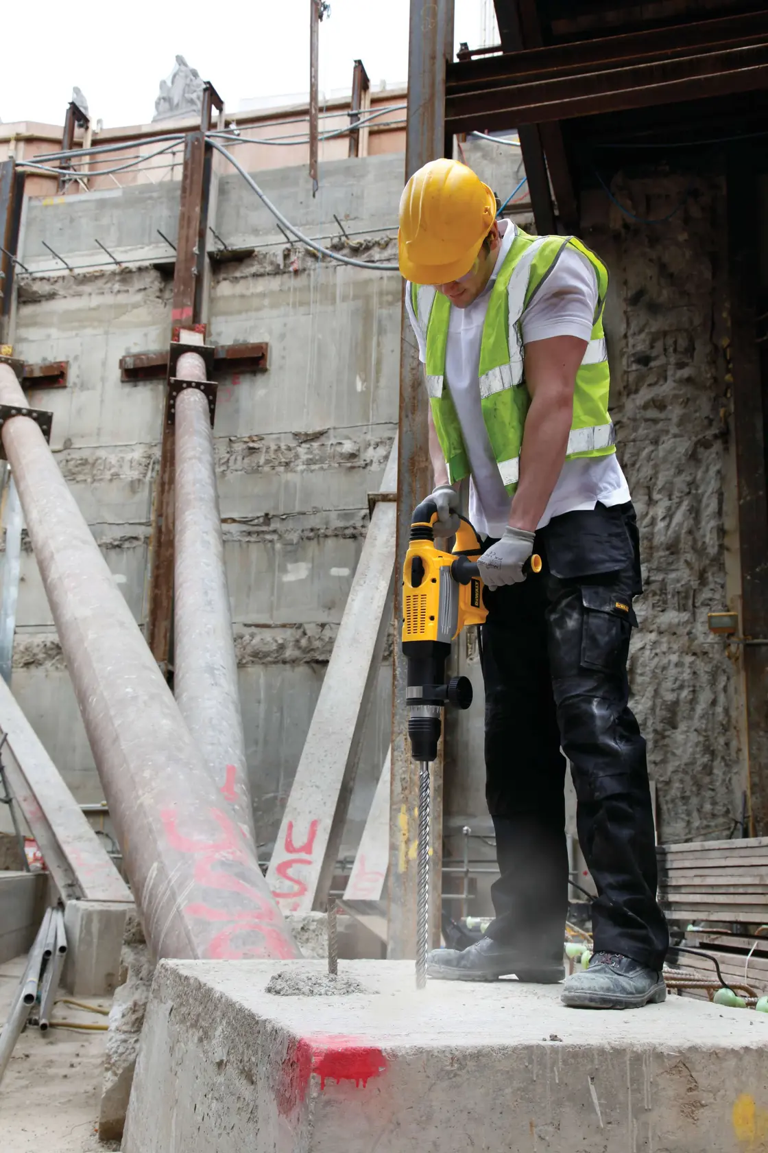 Worker uses yellow DEWALT drill on concrete at construction site. Wearing safety gear. DT9450 SKU visible.
