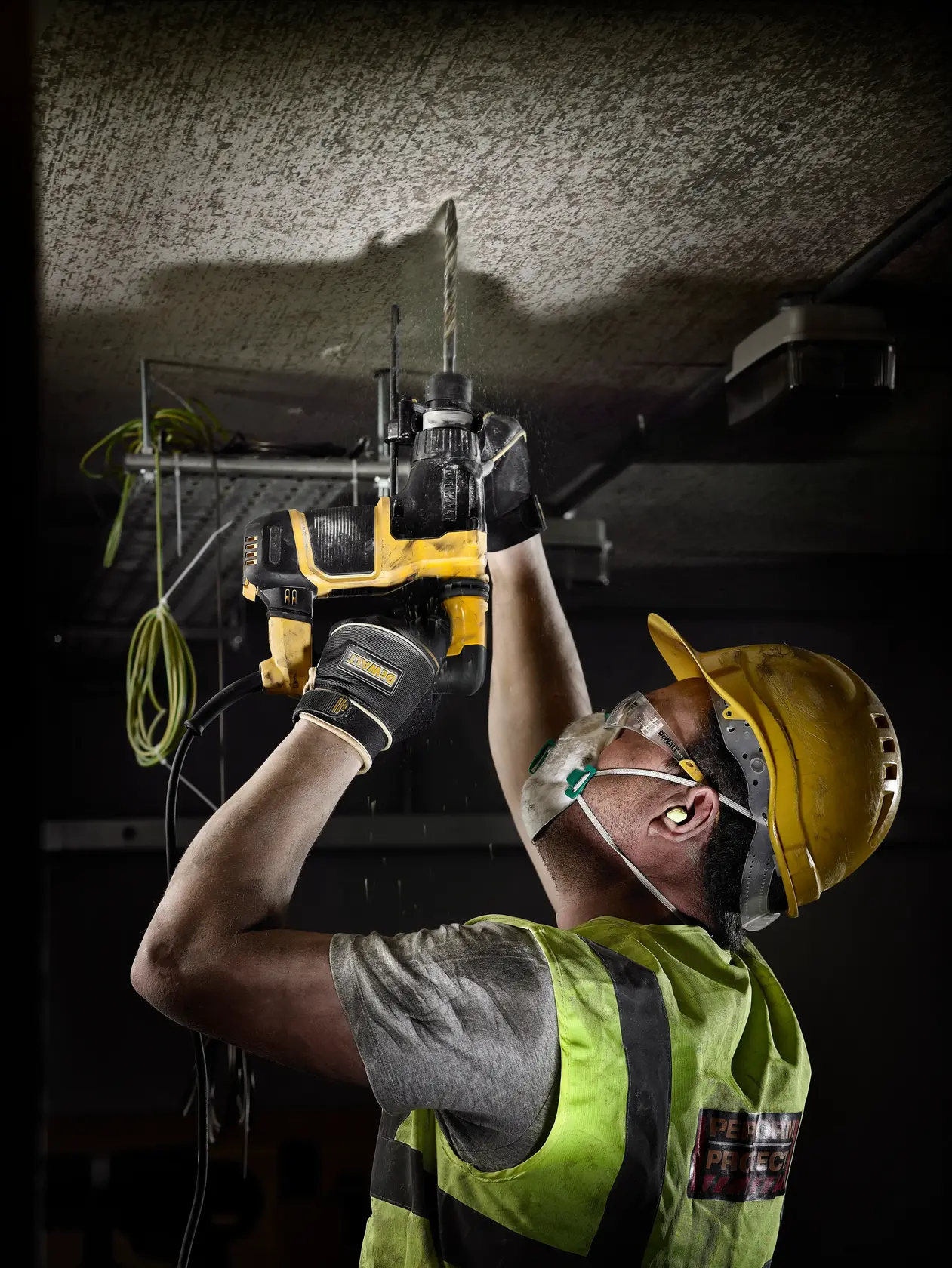 Worker uses a DEWALT drill to bore into a ceiling, wearing safety gear and a hard hat on a construction site.