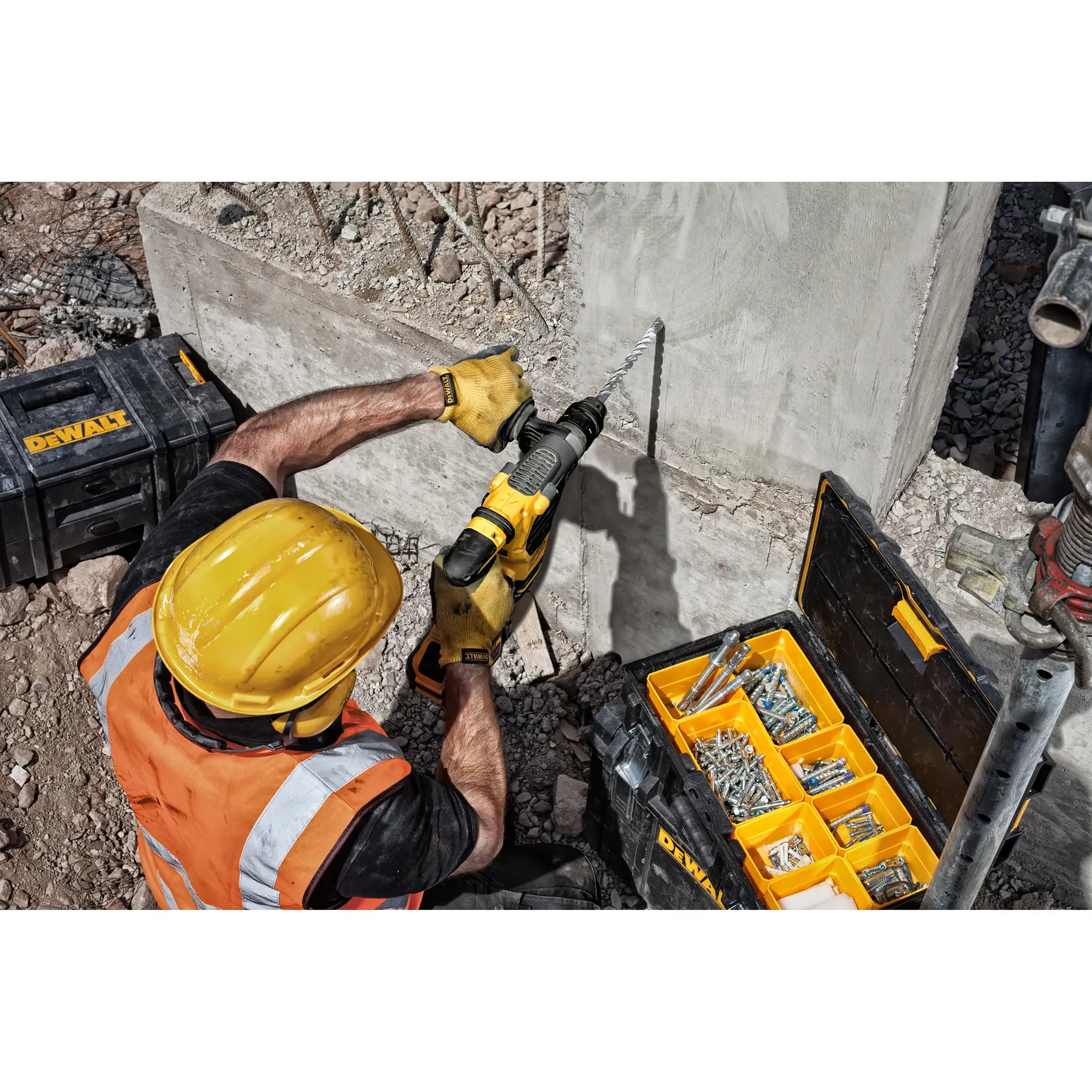 Worker with DEWALT drill making hole in concrete wall; toolboxes with screws and bolts are visible nearby.