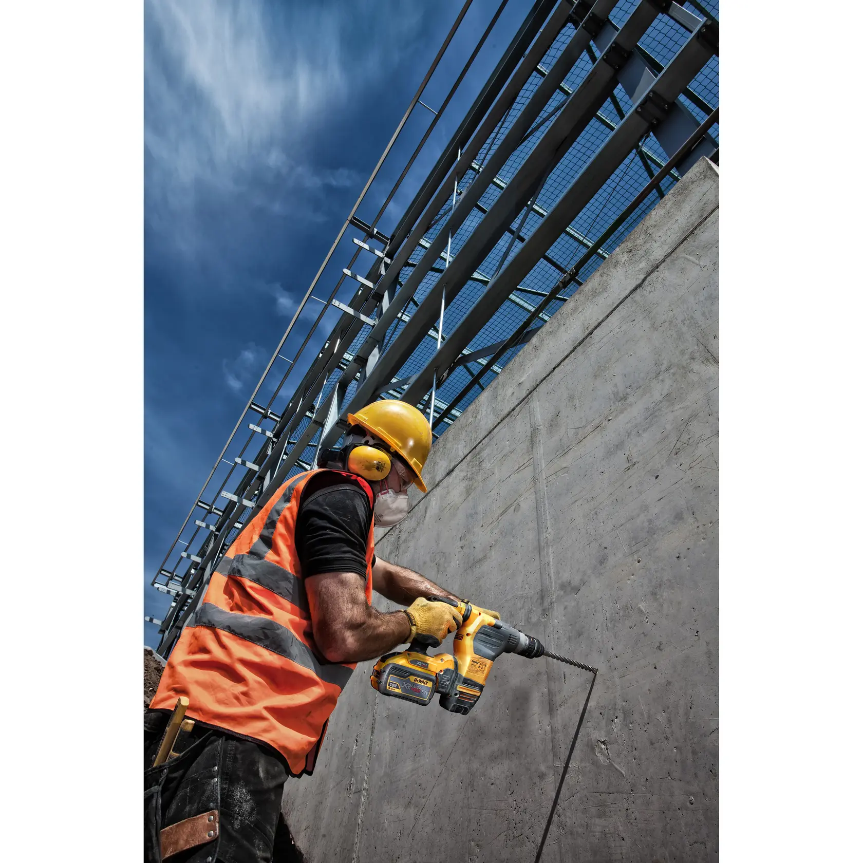 Worker uses a DEWALT drill on a concrete wall, wearing safety gear and an orange vest at a construction site.