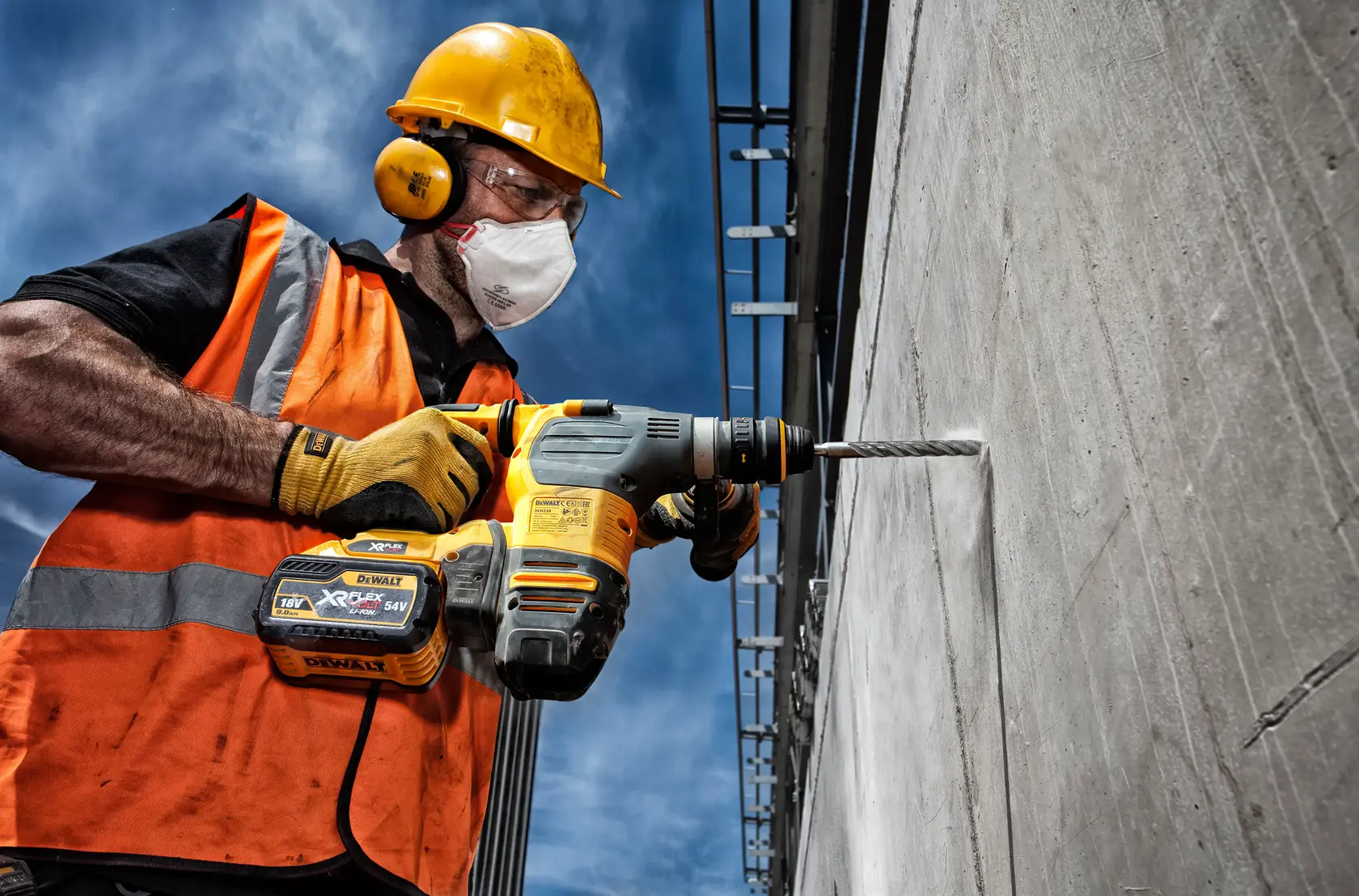 Construction worker drills into concrete using a DEWALT cordless drill. Worker wears safety gear and orange vest.
