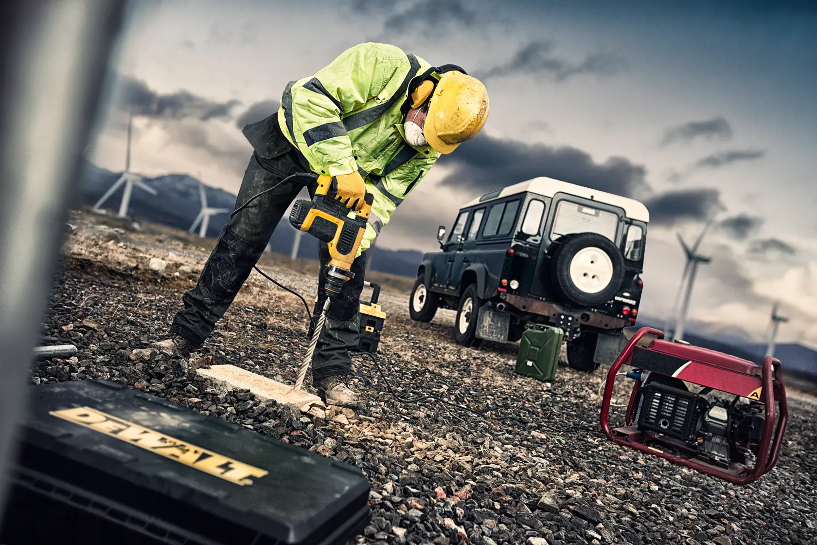 Worker drills with DEWALT tool on rocky ground, wearing safety gear, vehicle and wind turbines in the background.