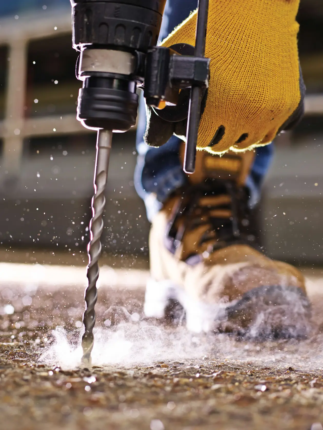 Close-up of a gloved hand using a DEWALT drill bit to drill into concrete, with dust and safety boots visible.