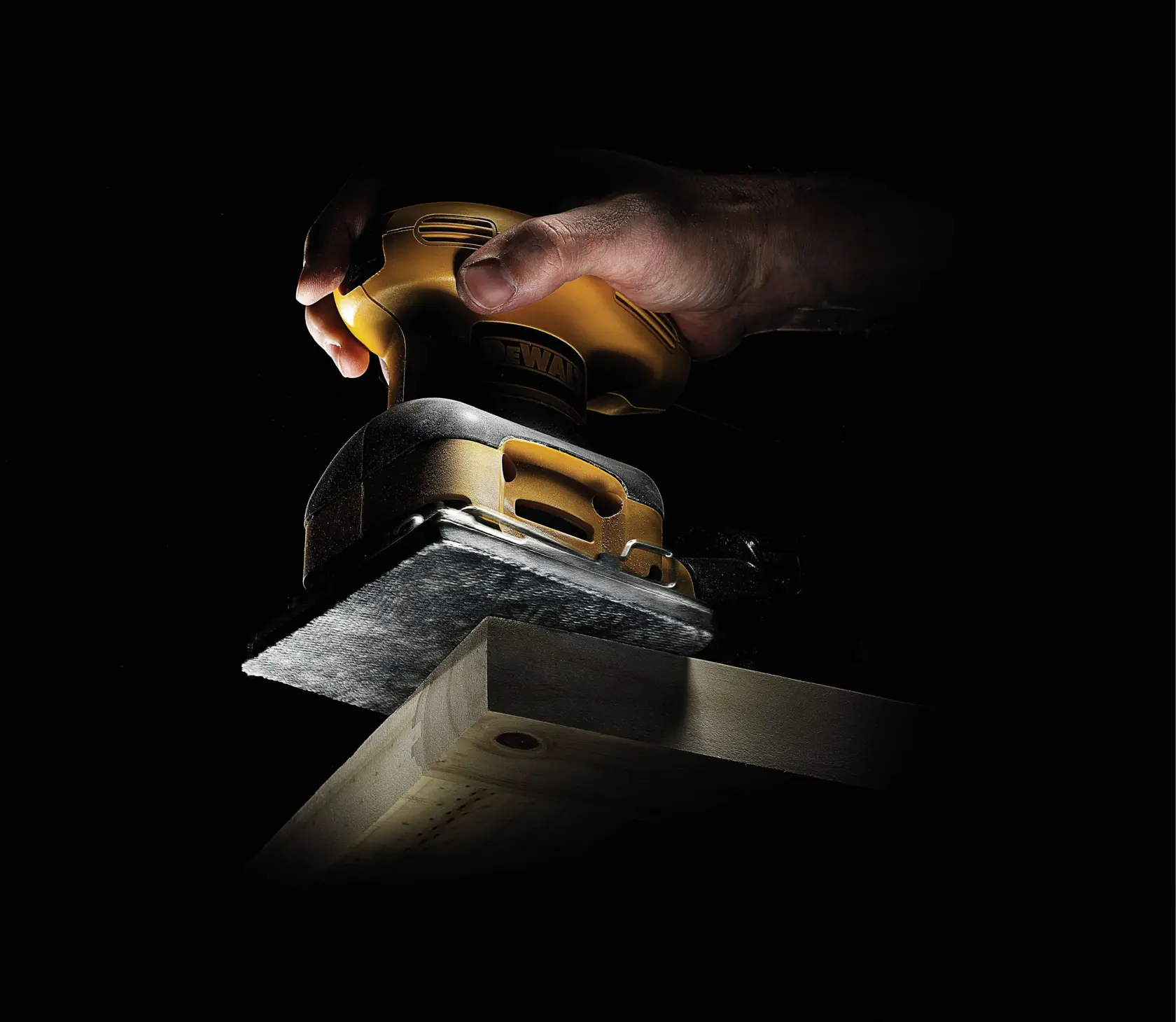Hand using a DEWALT sander to smooth the edge of a wooden plank against a dark background.