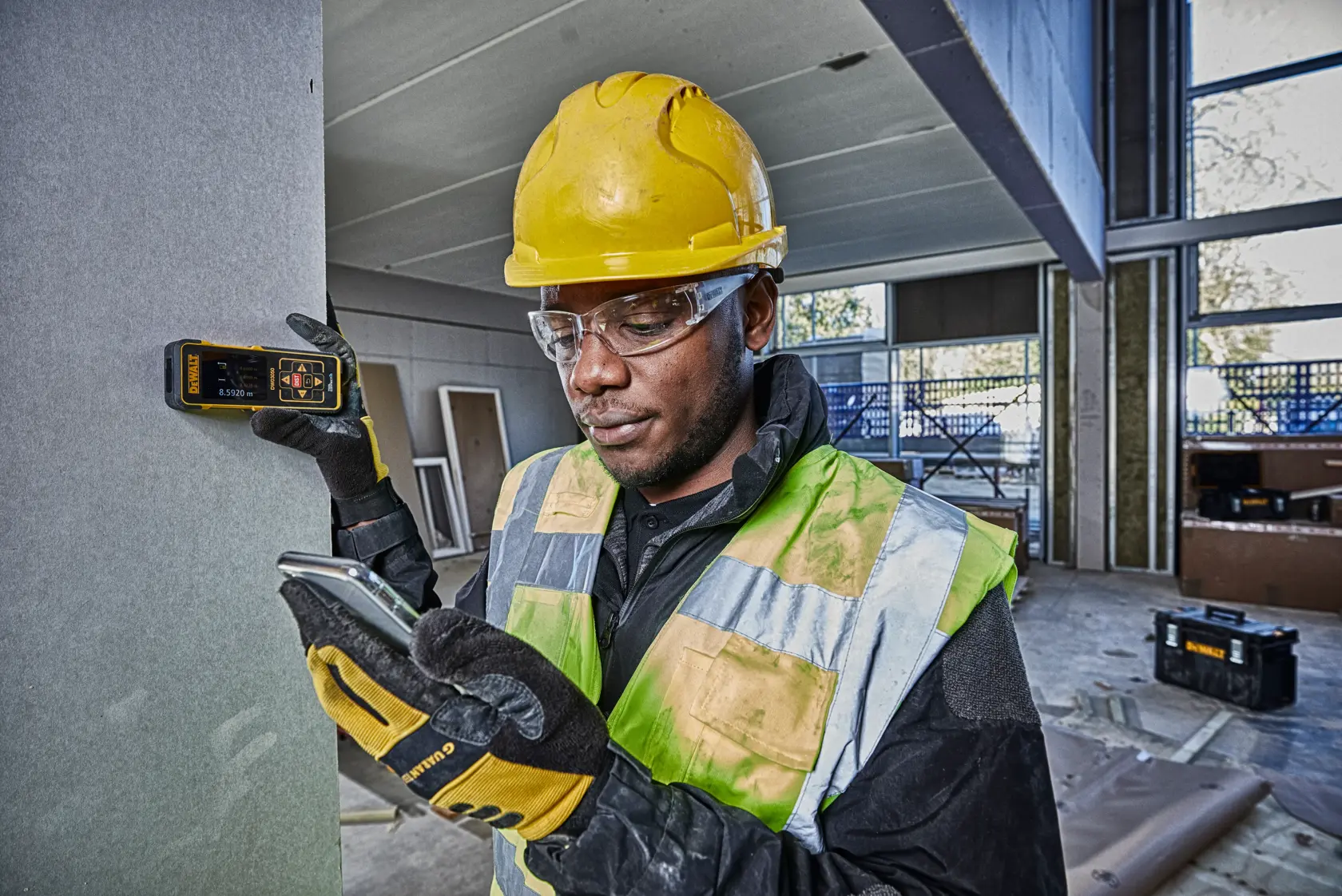 Worker uses DEWALT DW03050 laser distance measurer on construction site, wearing helmet and safety vest.