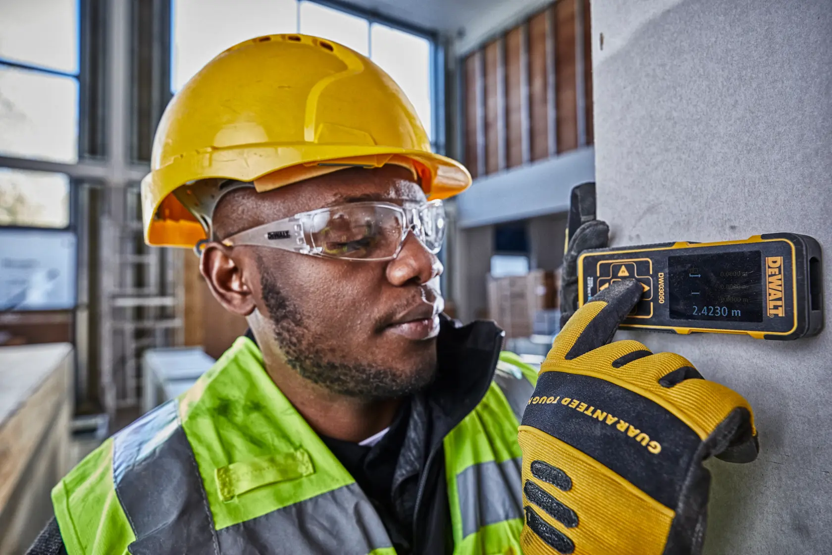Worker uses DEWALT DW03050 laser distance meter on construction site, wearing safety gear and gloves.