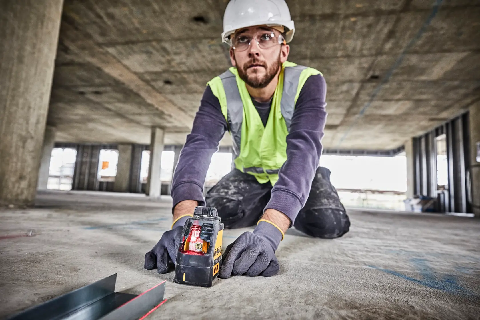 Worker using a DEWALT DW088K laser level on a construction site floor, wearing gloves and safety gear.
