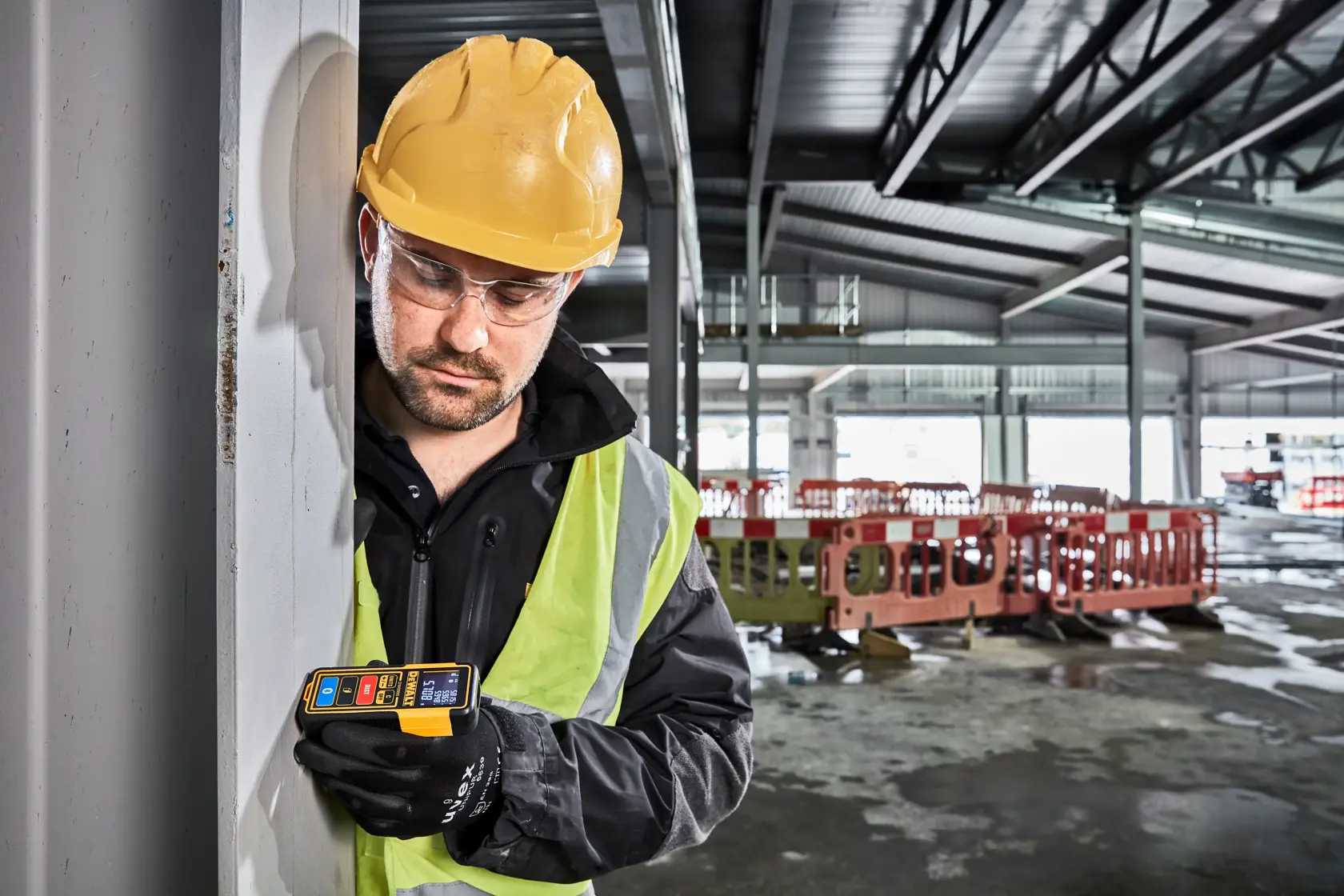 Worker in safety gear uses a DEWALT DW099S laser measure at a construction site indoors.