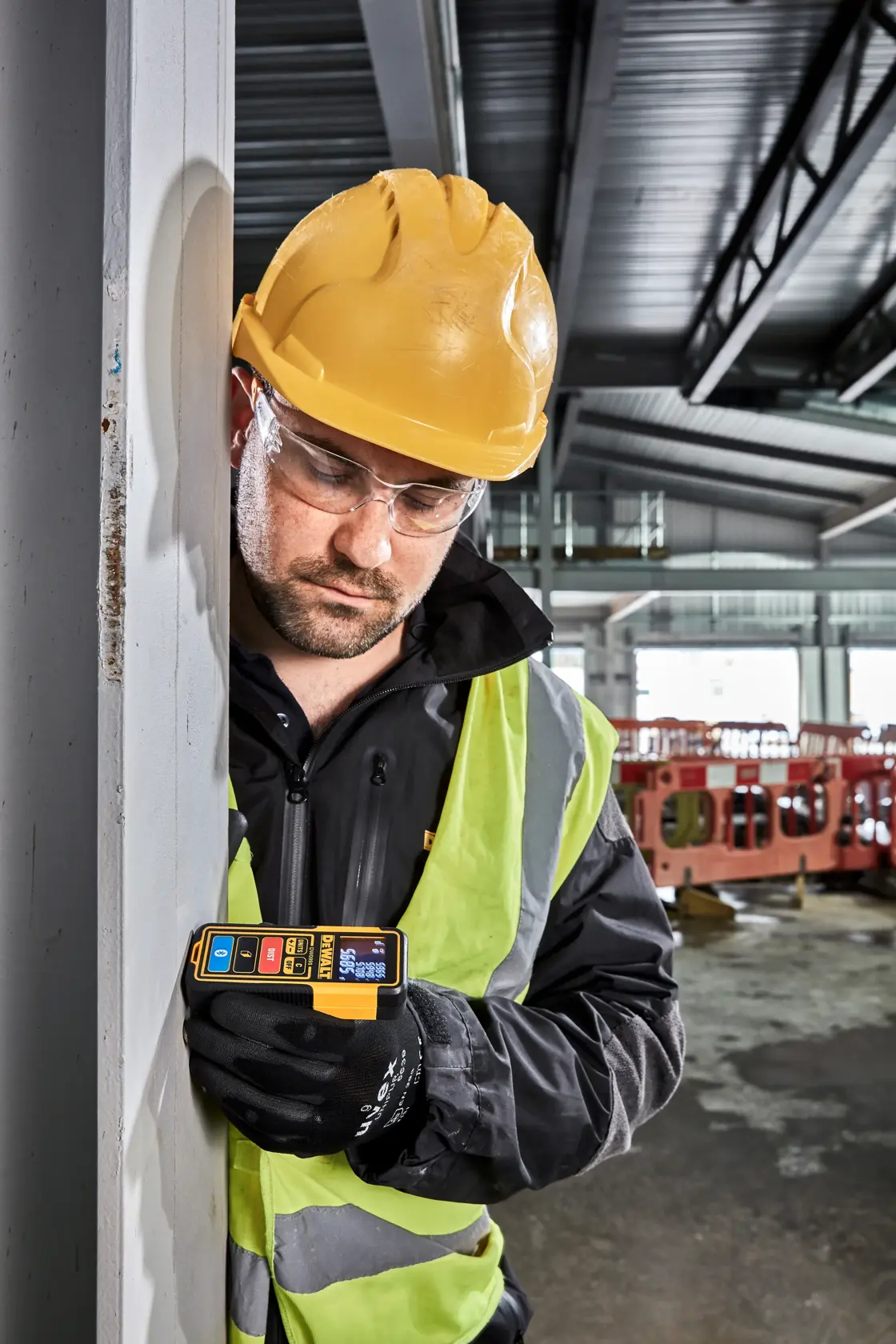 Worker uses DEWALT DW099S laser distance measurer at a construction site, wearing safety vest and helmet.