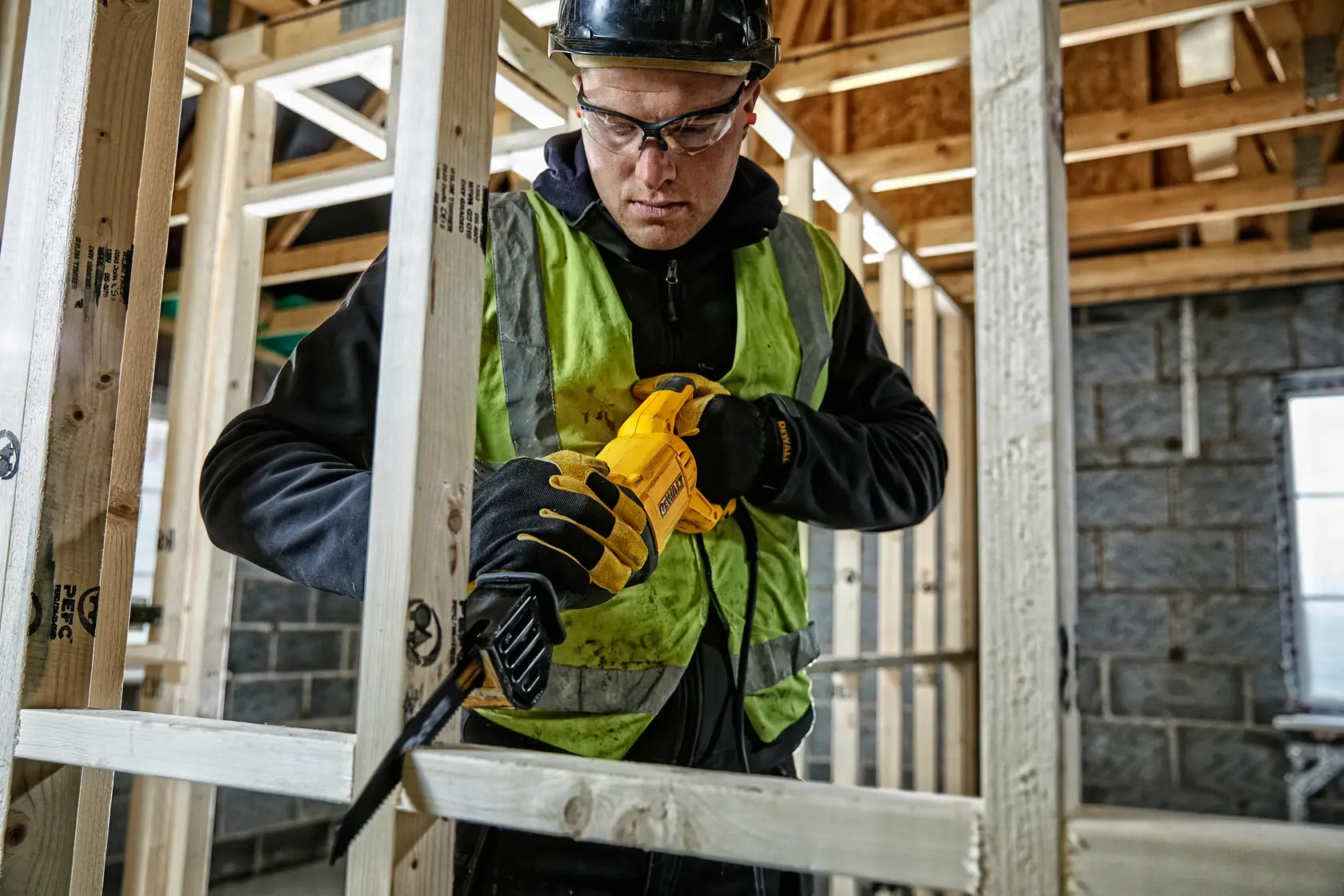 Worker uses a yellow DEWALT reciprocating saw to cut wood frame at a construction site, wearing safety gear.