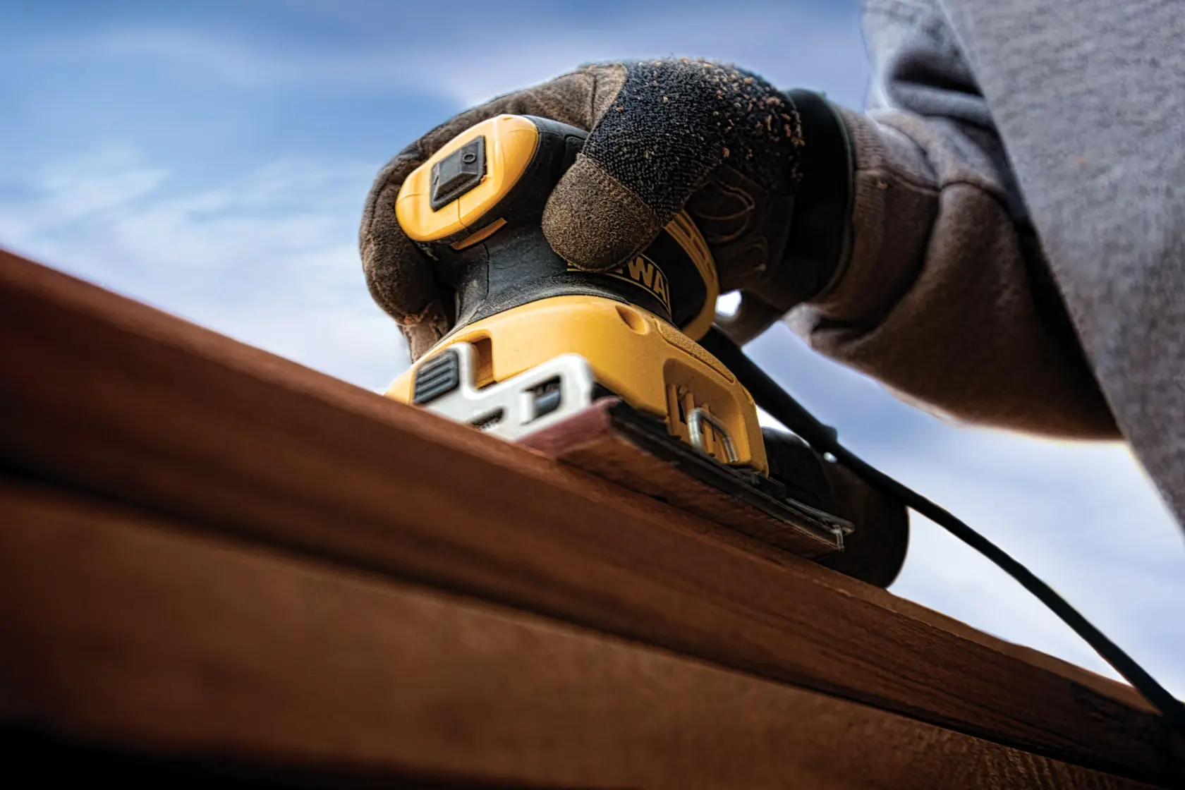 A close-up view of a gloved hand operating a yellow and black DEWALT DWE6411 electric sander on a wooden surface outdoors.