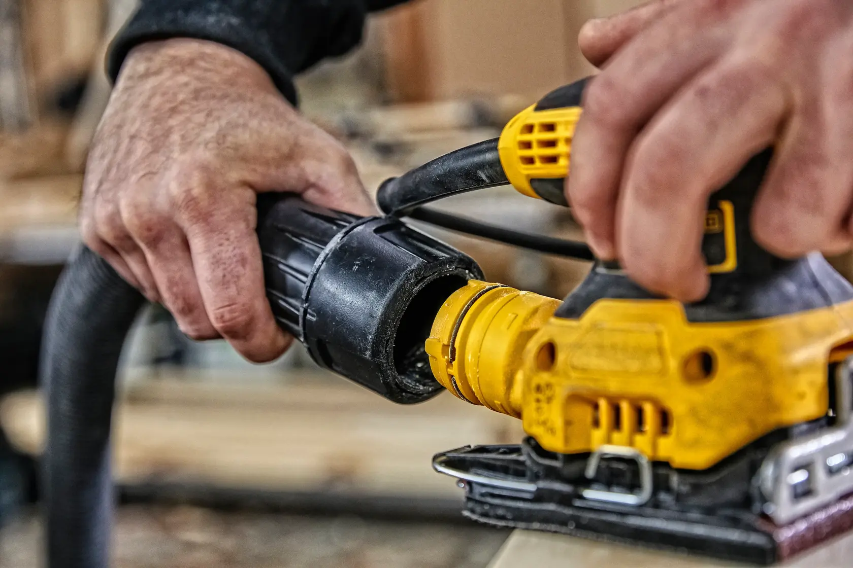 Hands connecting a hose to a yellow DEWALT power tool, used for woodworking or sanding.