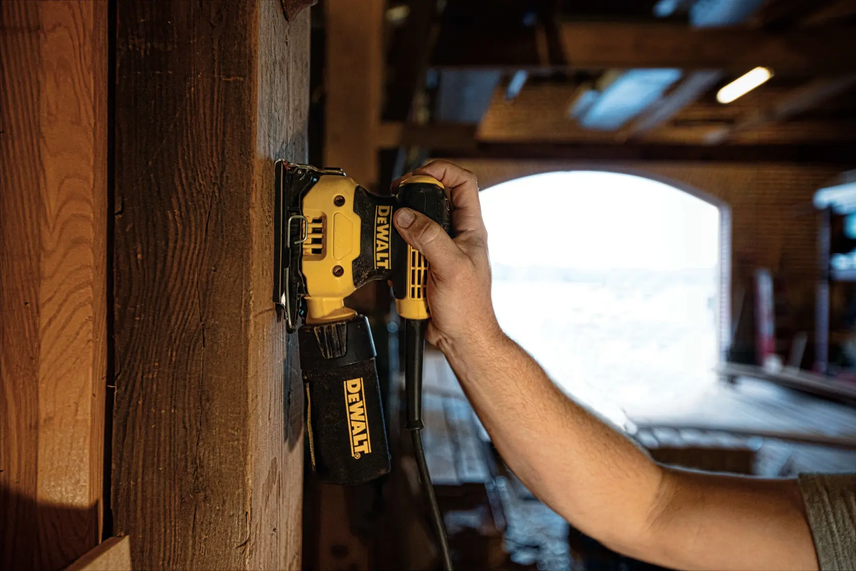 A person is using a DEWALT DWE6411 electric sander on a wooden surface inside a workshop, with the sander held vertically and dust bag attached.