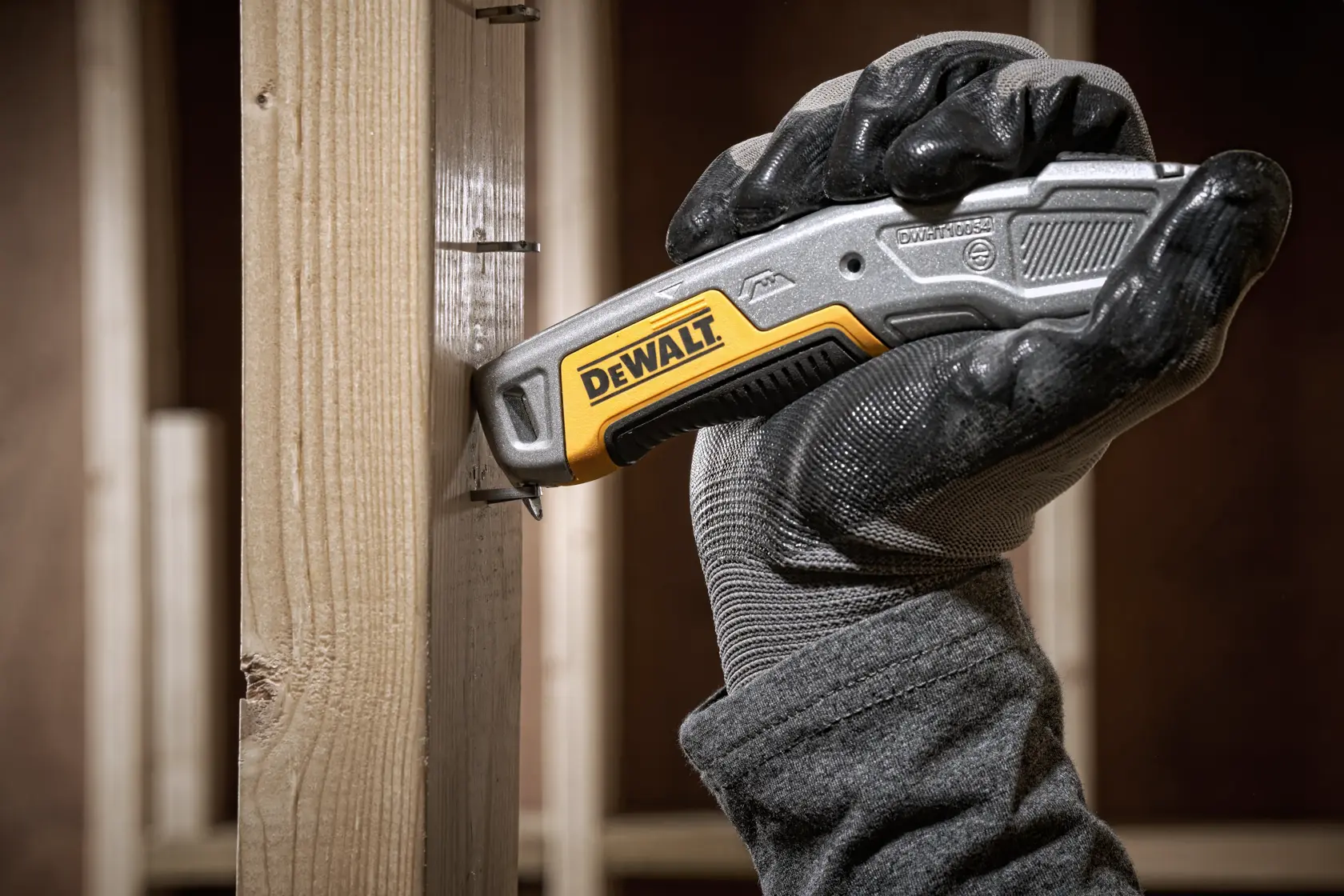 Gloved hand using a DEWALT utility knife against a wooden beam at a construction site.