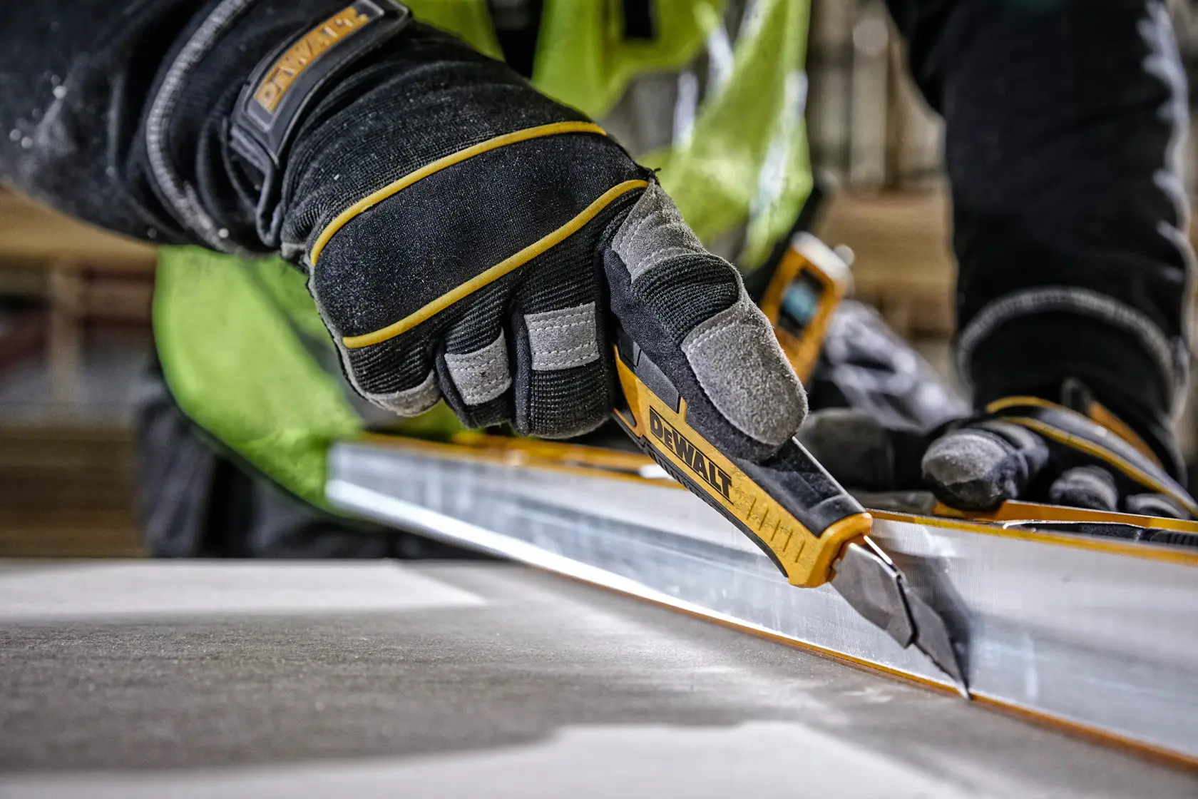 Gloved hand using DEWALT utility knife to cut material along a metal straight edge at a construction site.