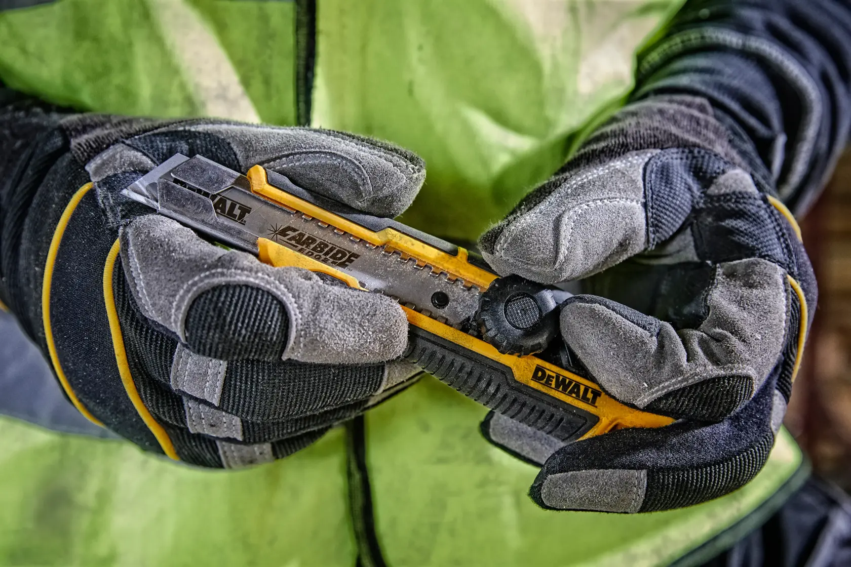 Gloved hands holding a yellow DEWALT utility knife with carbide blade against a high-visibility jacket background.