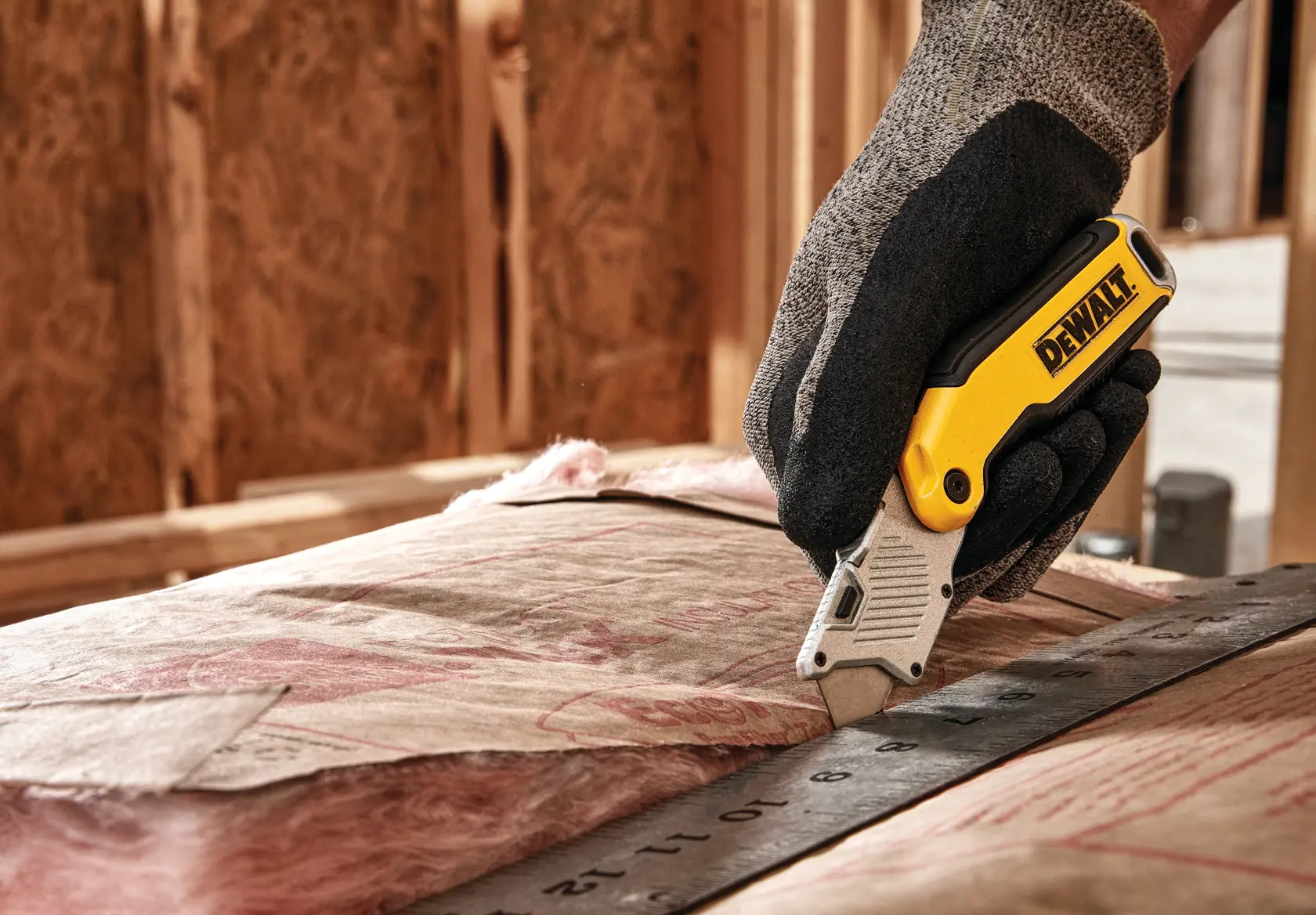 A gloved hand uses a DEWALT utility knife to cut insulation material along a metal ruler at a construction site.