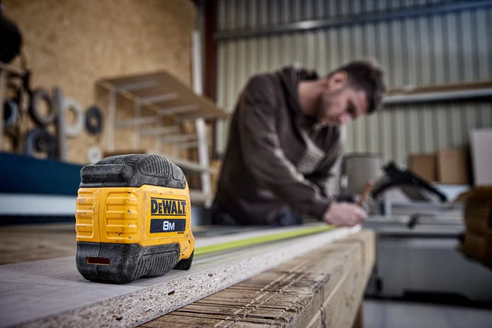 DEWALT 8M tape measure on a wood surface, person measuring in the background inside a workshop.