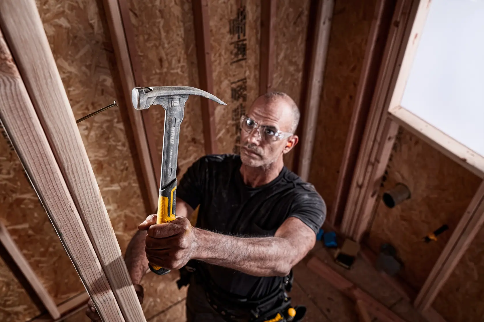 A person using a DEWALT hammer (model DWHT51135-0) to drive a nail into a wooden frame inside a partially constructed room. The hammer has a yellow and metal handle.