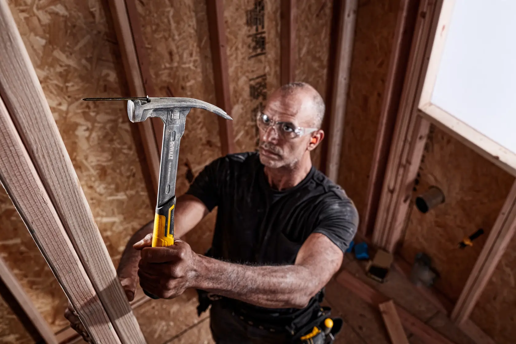 A person using a DEWALT hammer (DWHT51135-0) to work on a wooden frame inside a construction site. The hammer has a yellow and black handle, and the person is wearing a tool belt.