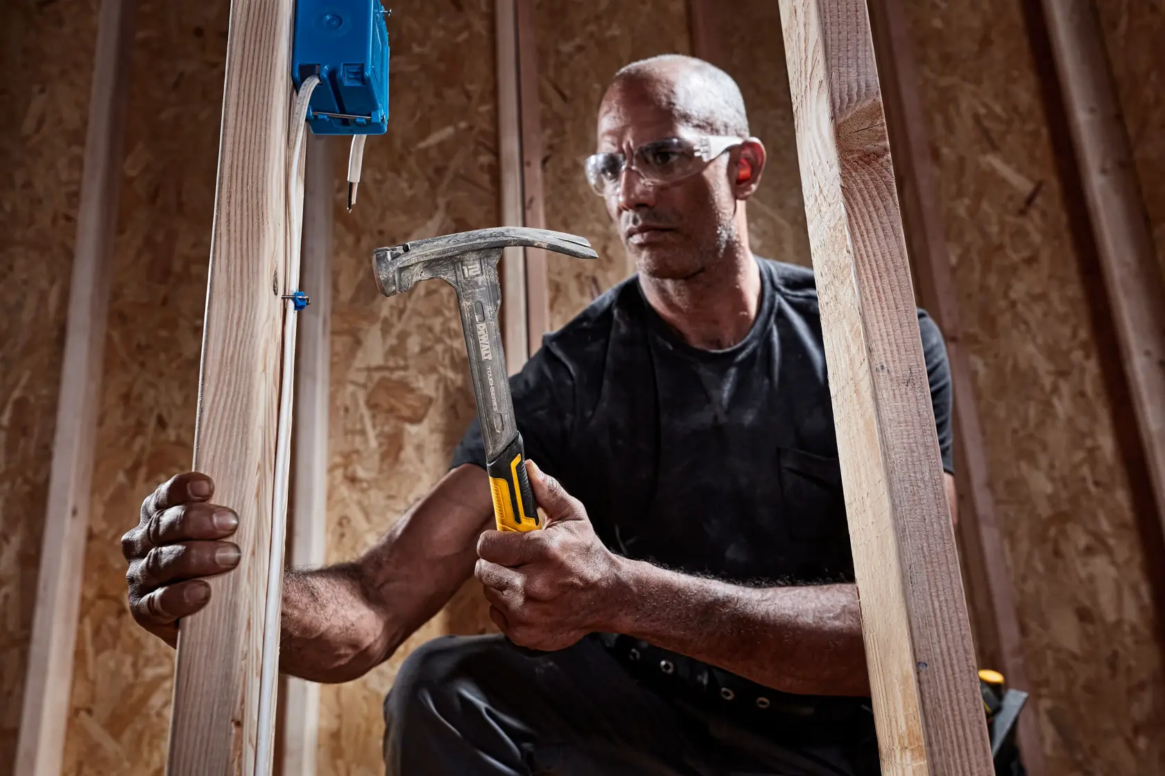 A person holding a DEWALT claw hammer with a yellow and black handle, working on a wooden construction frame. Their face is blurred and they appear to be installing an electrical box onto wooden beams.