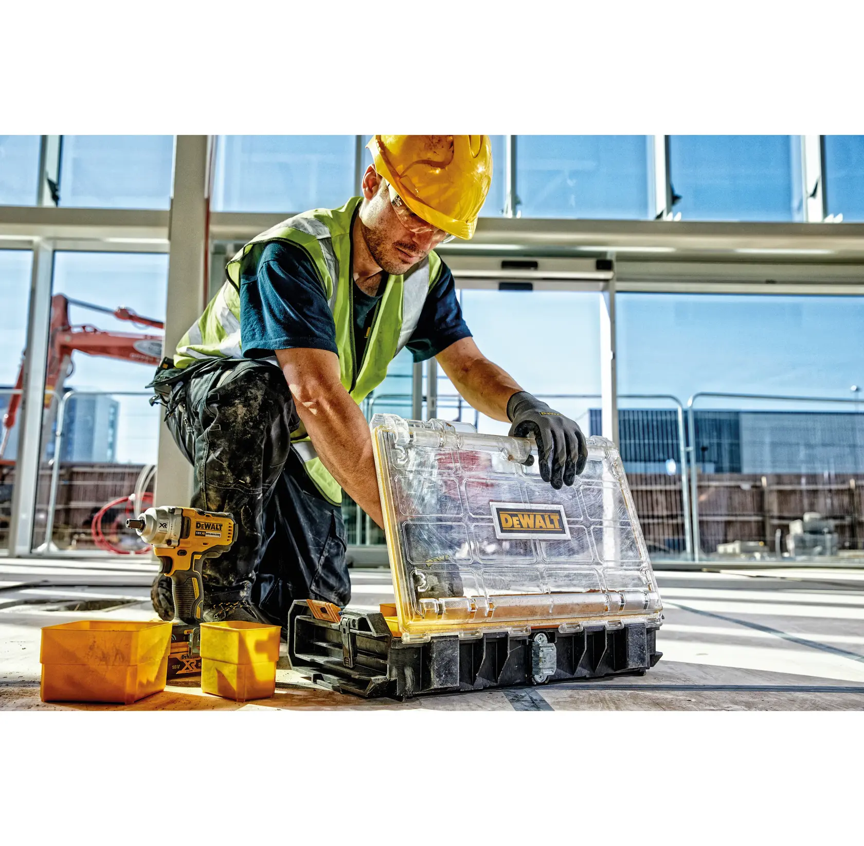 Worker on site with DEWALT tool case, drill, and containers near a window. Safety gear is worn.