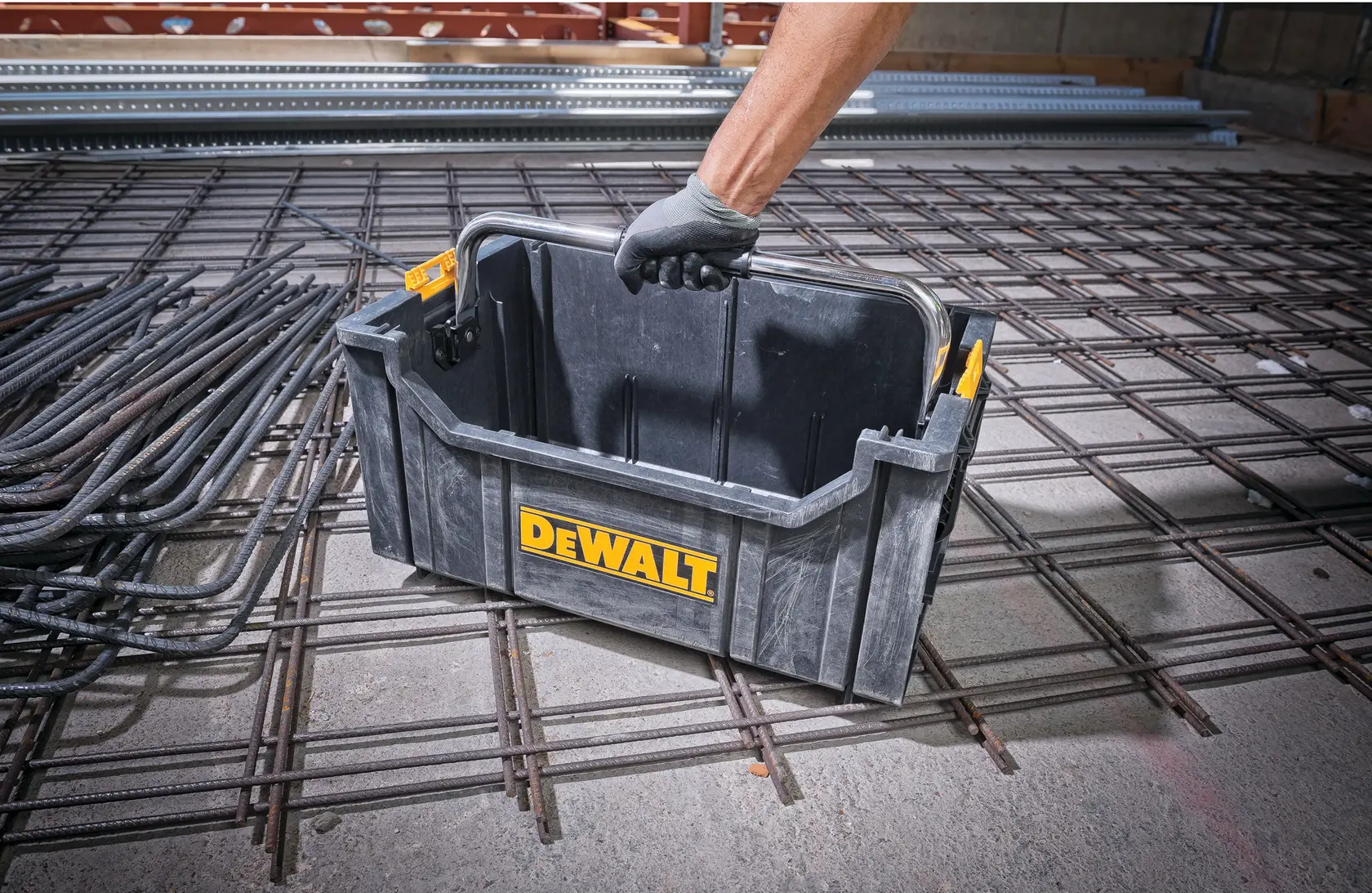 Person lifts a DEWALT plastic tool box with a handle, placed on a metal grid in a construction site.