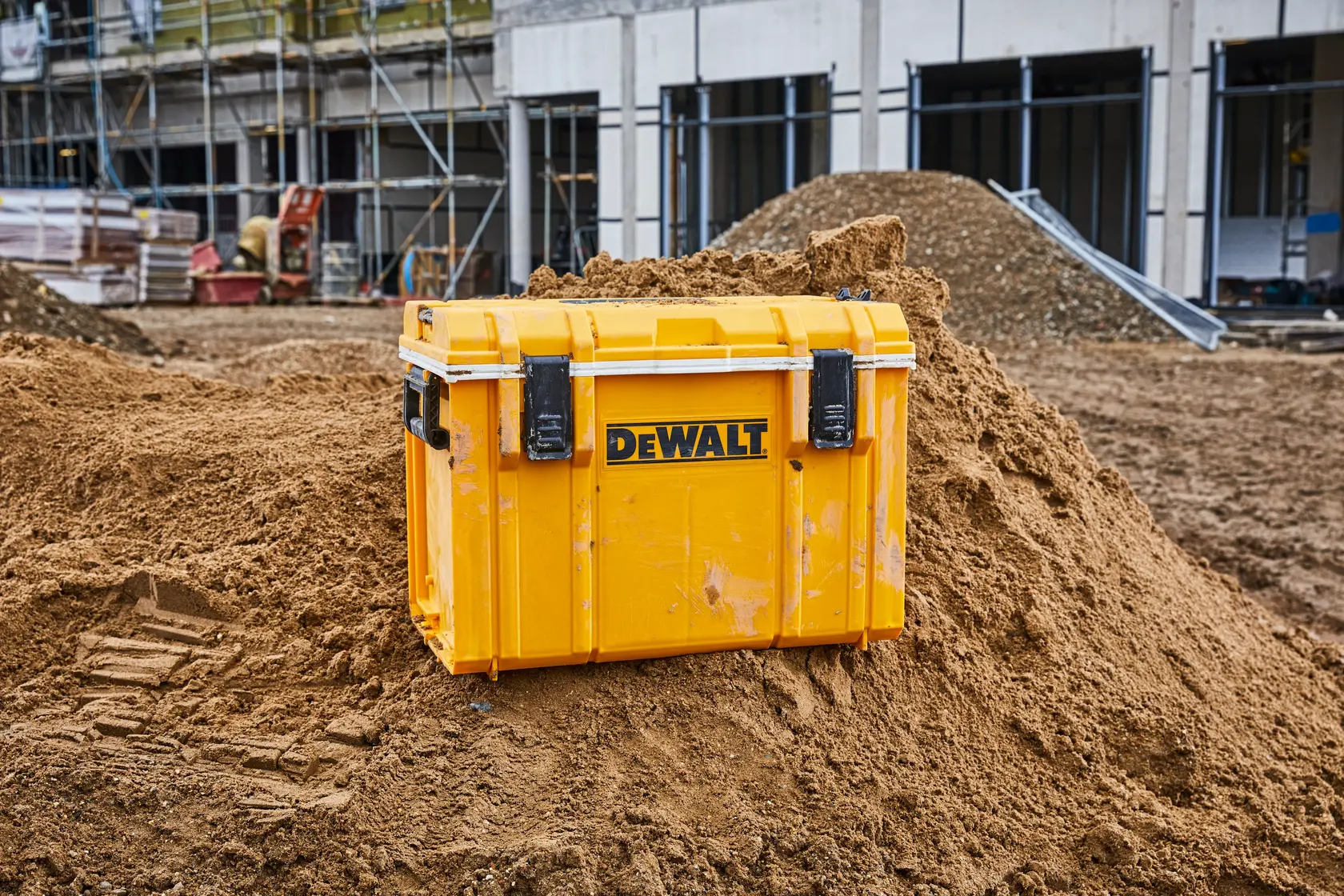 Yellow DEWALT tool box on a dirt mound at a construction site with building scaffolding in the background.