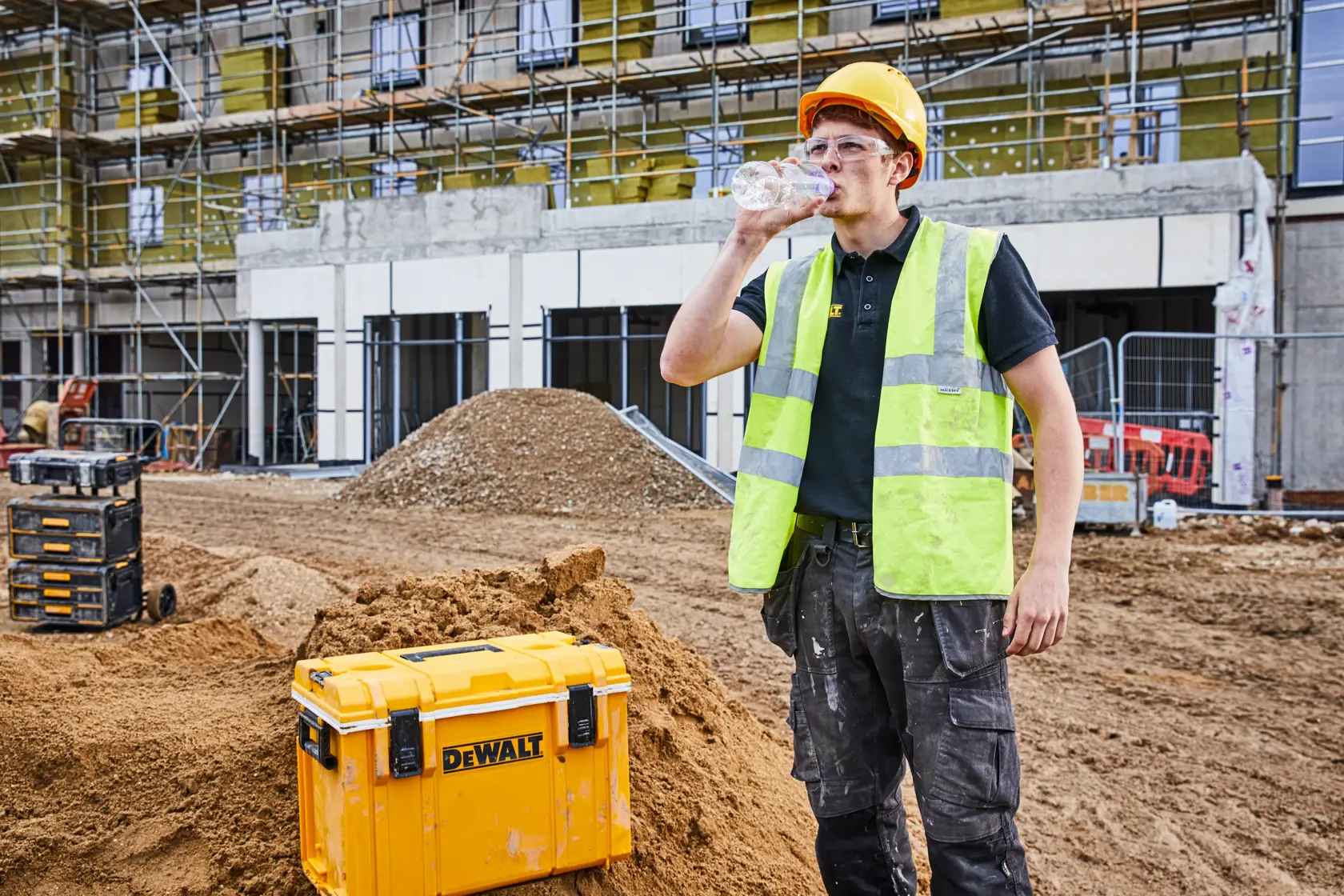 DEWALT storage box on a construction site, worker in safety vest and helmet drinks water.