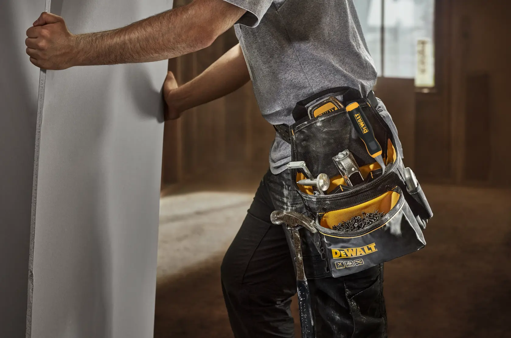 A person wearing a DEWALT tool belt holds a large panel. The tool belt contains various hand tools, including a hammer, screwdrivers, and fasteners. The scene is set indoors, suggesting a construction or installation environment.