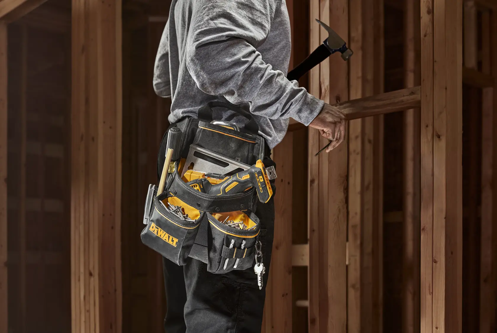 A person wearing a DEWALT tool belt loaded with various hand tools, working in a wooden structure. The tool belt has multiple pockets and compartments for organized tool storage.