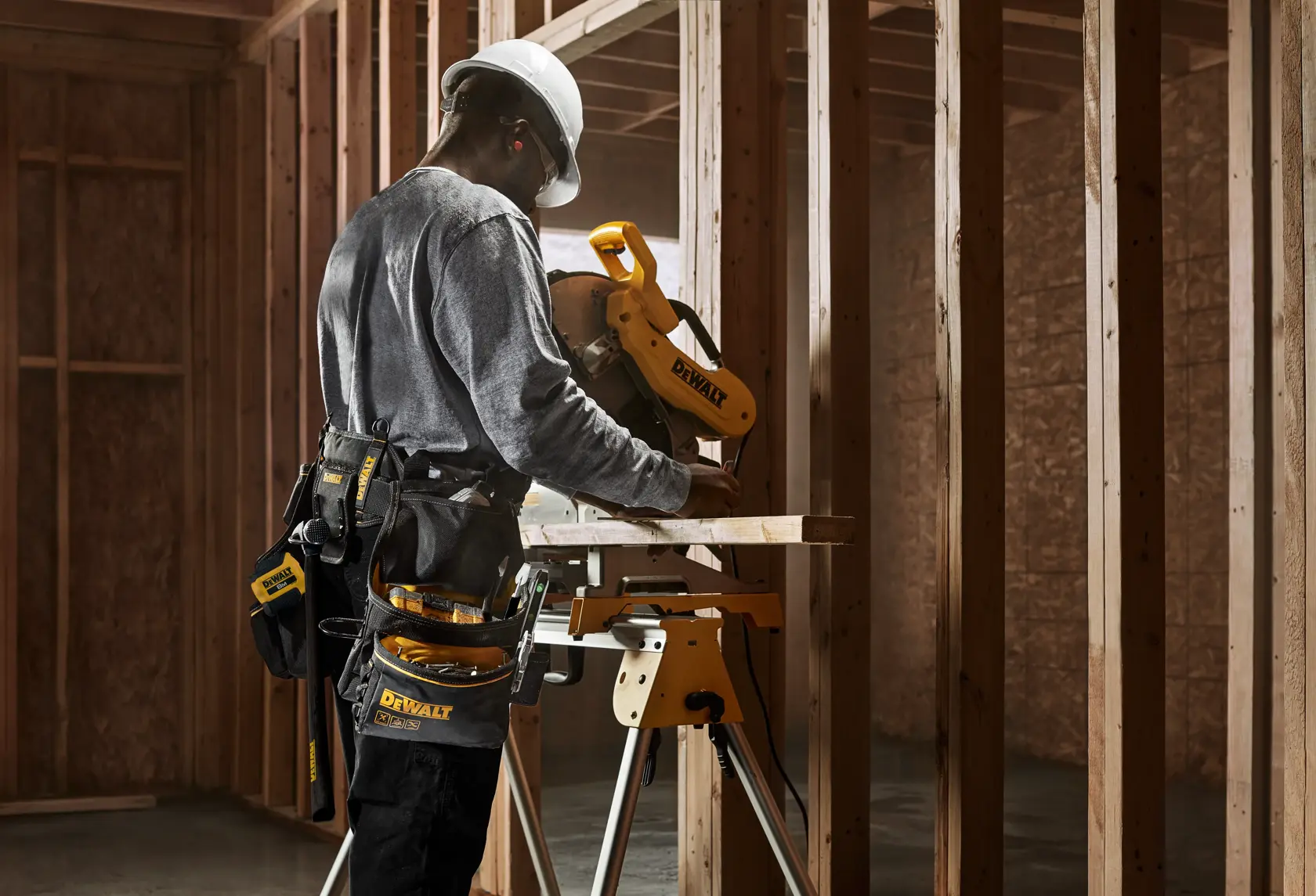 Person wearing a hard hat and tool belt operating a DEWALT miter saw in a wooden frame construction site.