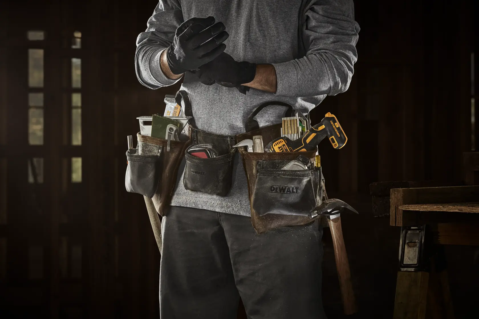 A person wearing a DEWALT tool belt full of tools, including a measuring tape, screwdrivers, pencils, and a yellow and black cordless drill, standing in a workshop and putting on black work gloves.