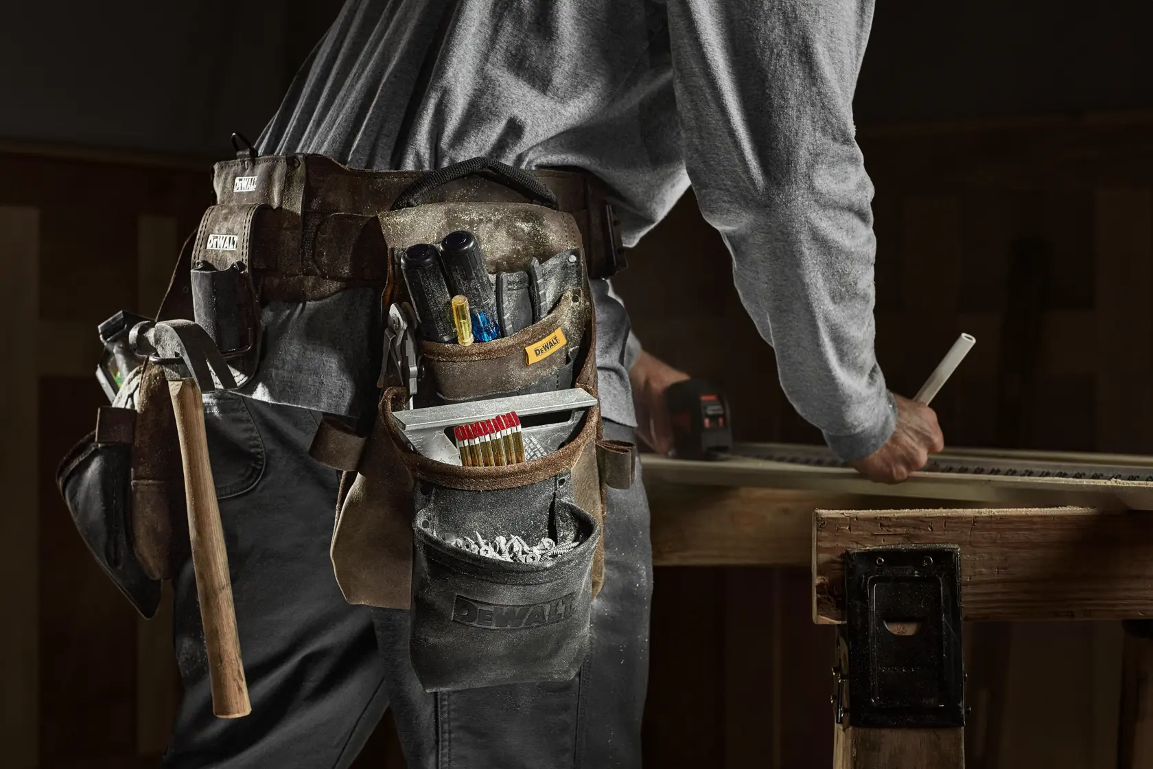 A close-up of a person wearing a DEWALT tool belt loaded with various tools such as a hammer, pencils, and screwdrivers. The person is working at a wooden table and marking a piece of wood.