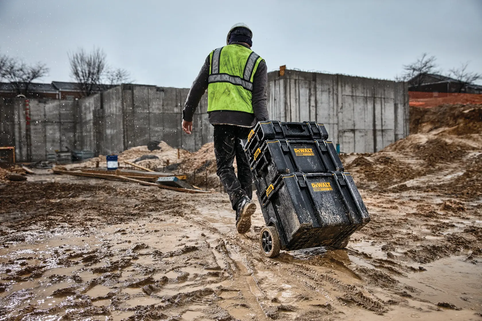 Worker pulls DEWALT toolboxes on wheels across a muddy construction site while wearing a safety vest and helmet.
