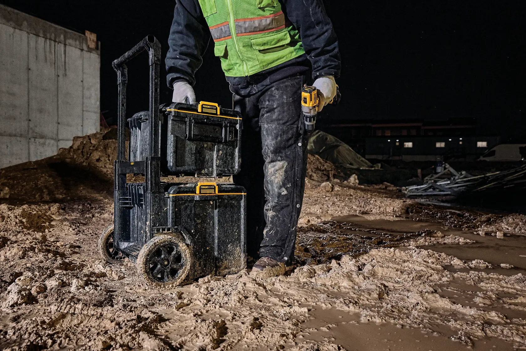 Worker with DEWALT tool boxes on wheels in muddy construction site at night.