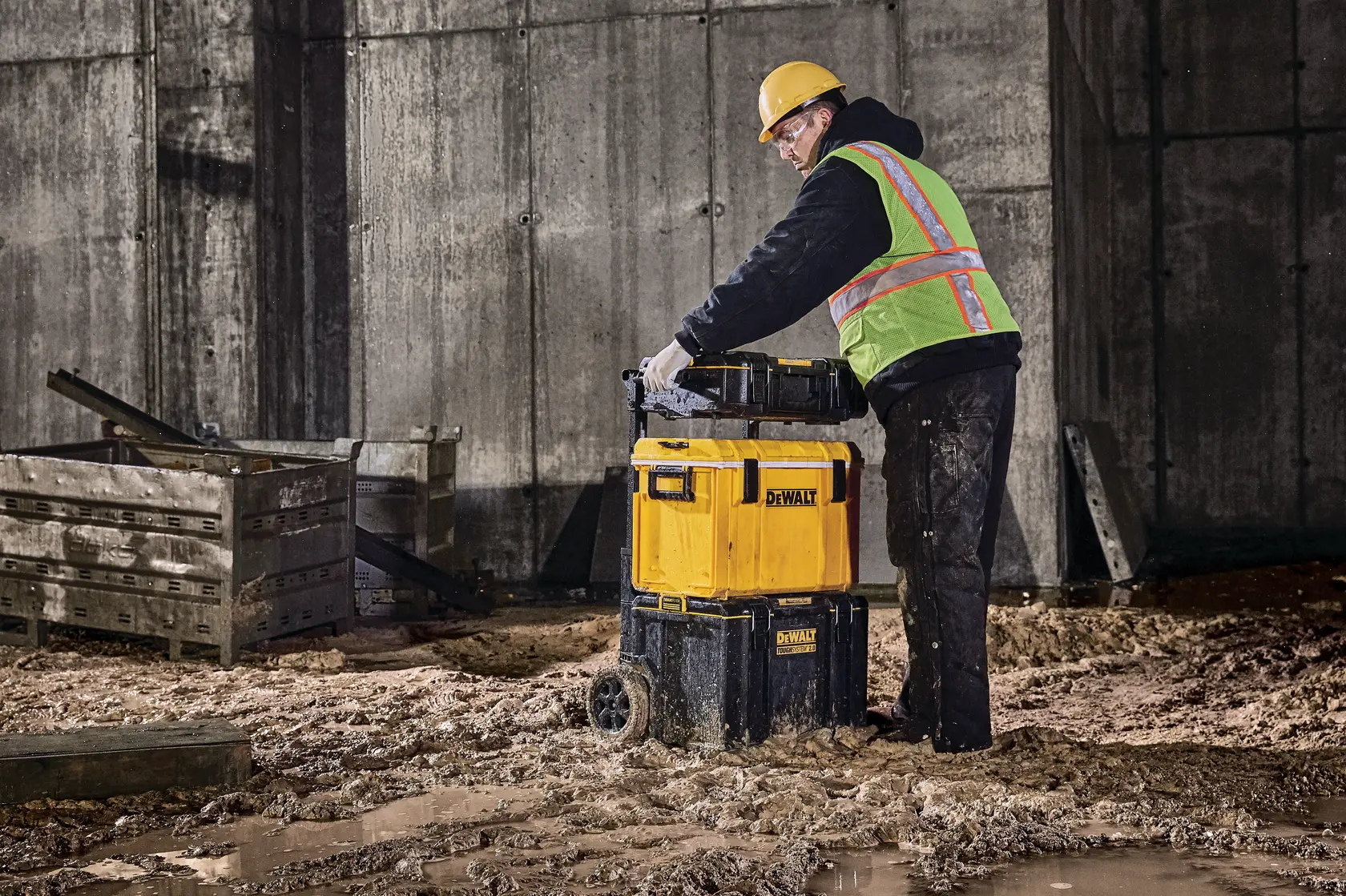 Worker moves DEWALT wheeled toolbox on muddy construction site near wooden crates and concrete wall.