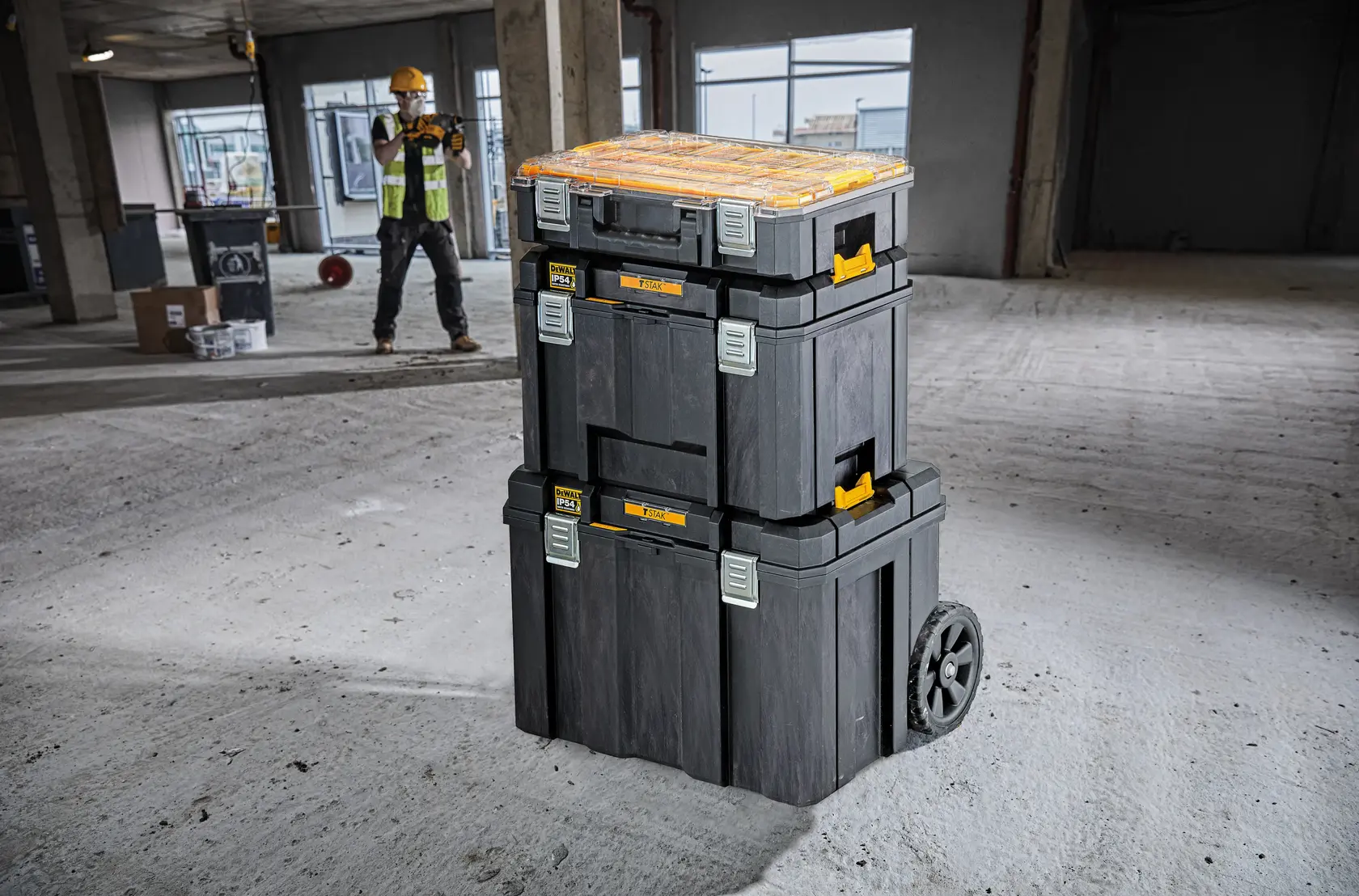 Stacked DEWALT tool boxes with wheels in a construction area, worker in background wearing safety gear.