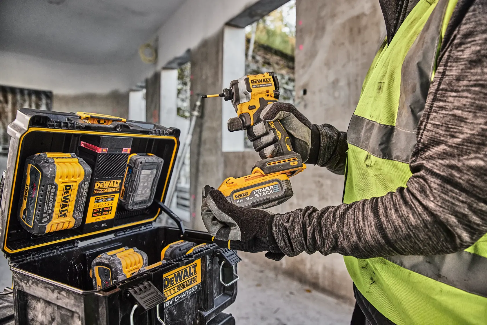 Person holding a DEWALT power tool and battery, with DEWALT tool case and batteries visible at a construction site.